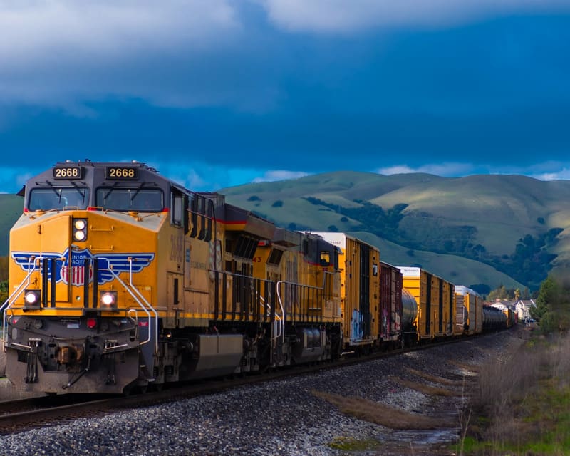 Yellow freight locomotive pulling cargo cars through a mountainous rail corridor.